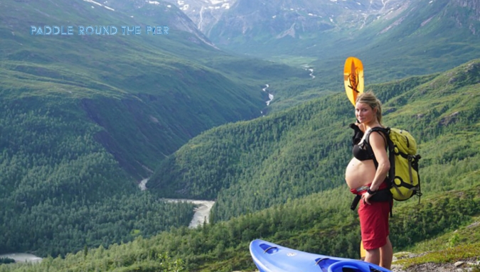 Pregnant woman with a kayak and gear overlooking a river in a mountainous forest, highlighting kayaking during pregnancy.